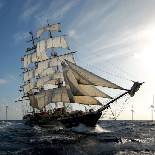 Large sailing ship on rough ocean water beneath a bright blue sky with wind turbines in the distance