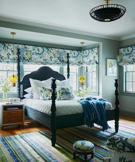 Sunlit bedroom with a black four-poster bed, striped rug, floral curtains, and blue bedding accents