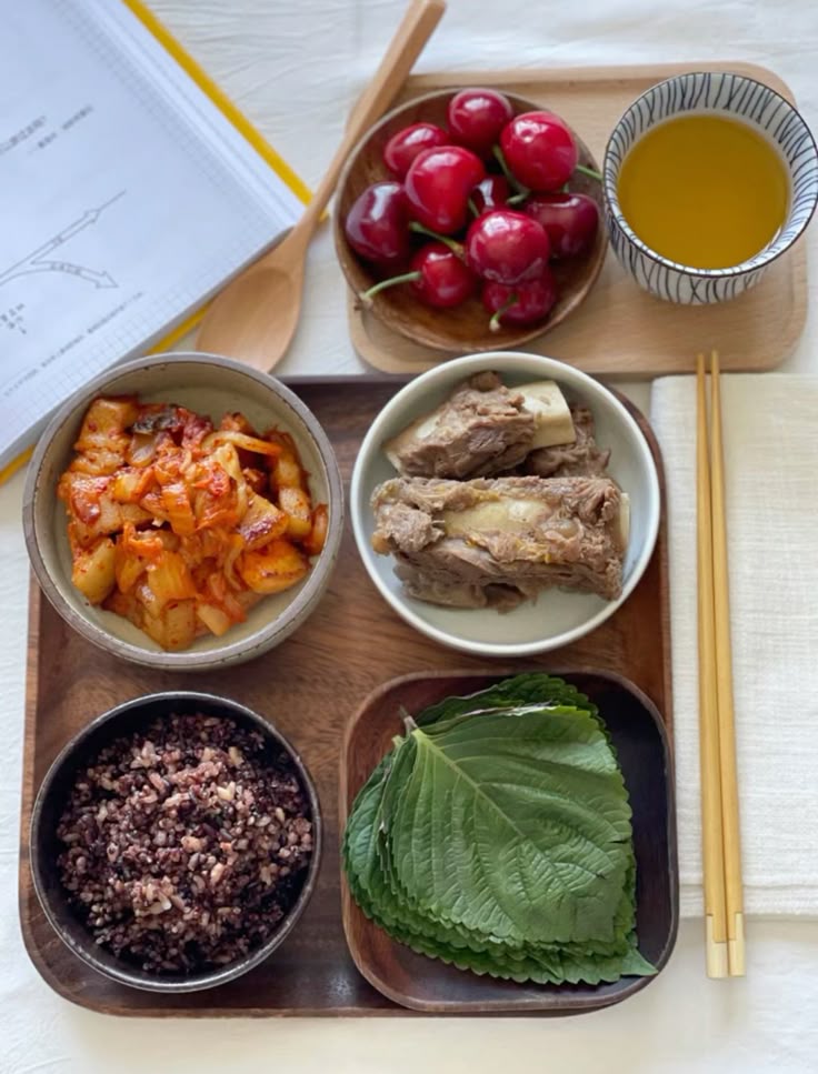 Wooden tray with Korean meal including kimchi, ribs, purple rice, perilla leaves, cherries, and tea