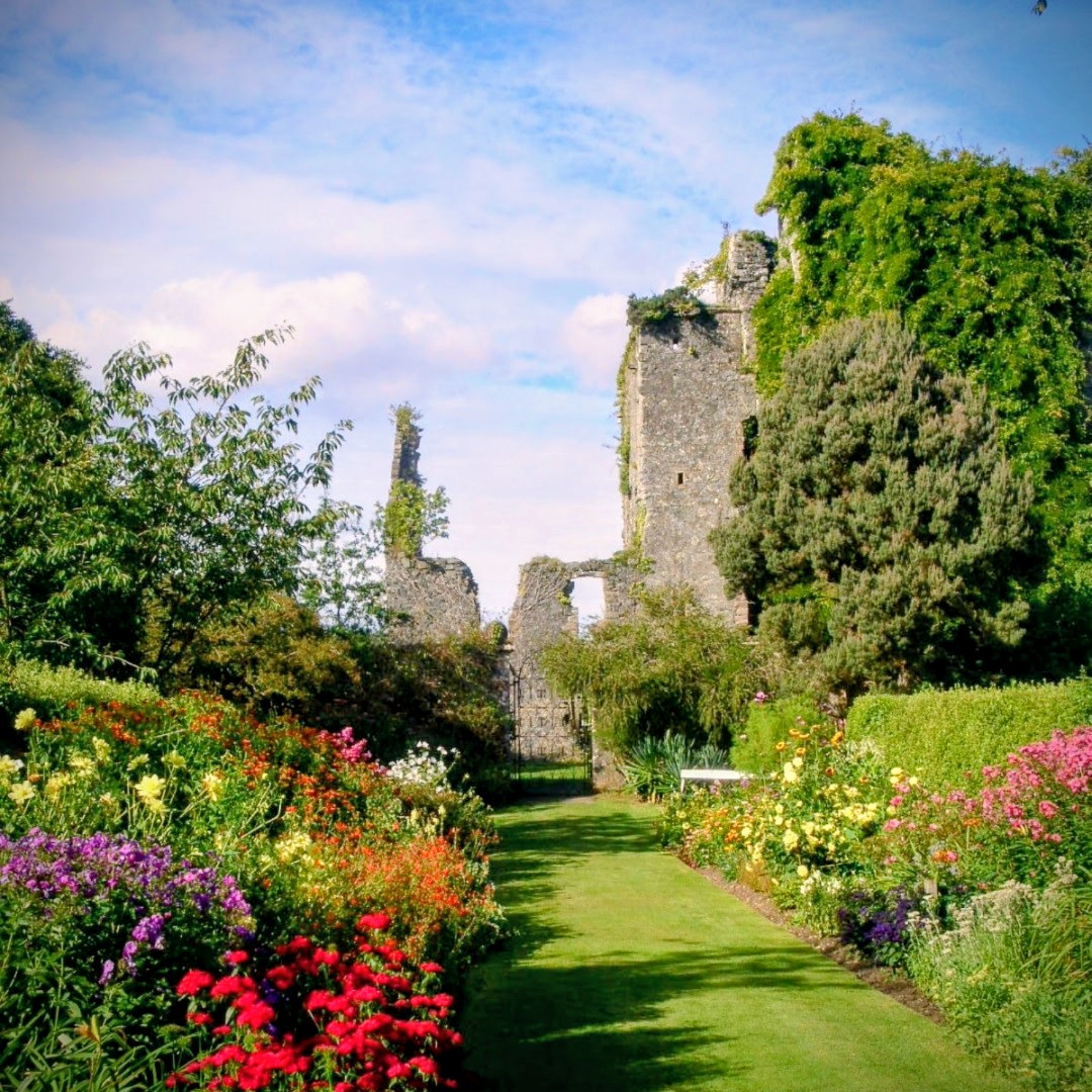 Stone castle ruins with ivy-covered towers in a vibrant garden with colorful flowers and manicured lawn