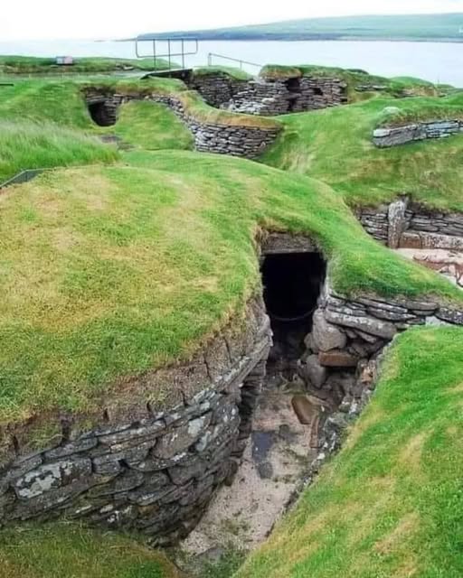 Sunken stone ruins of Skara Brae Neolithic village with grass-covered walls and a coastal bay in the background