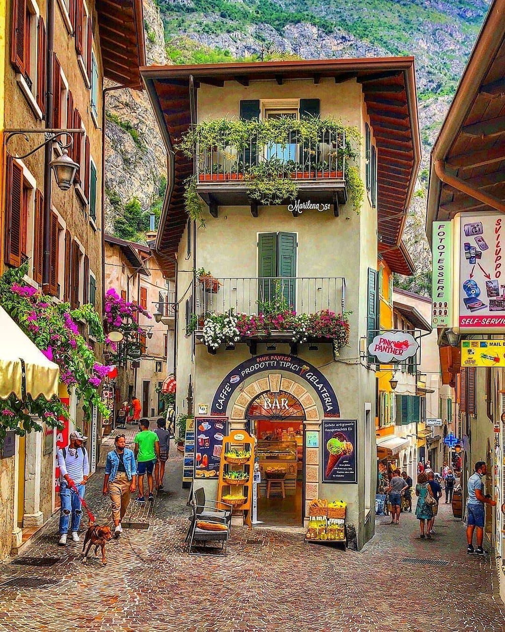 Picturesque Italian village street with colorful buildings, flowers, mountains, and tourists