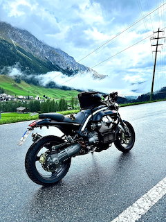 Black Moto Guzzi motorcycle on a wet road with mountains and clouds behind it