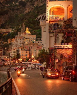 Curving coastal road with cars beside illuminated hillside village buildings at dusk