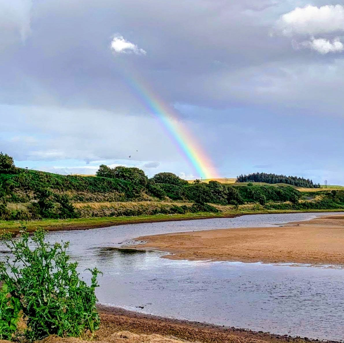 Colorful rainbow over a winding river with green hills and cloudy sky