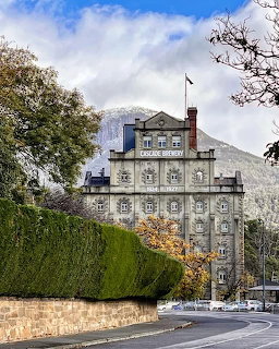 Historic multi-storey stone building with flag, framed by hedges and trees with a mountain behind