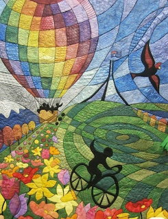 Textile quilt art showing a vibrant hot air balloon over a patchwork countryside with flowers and birds