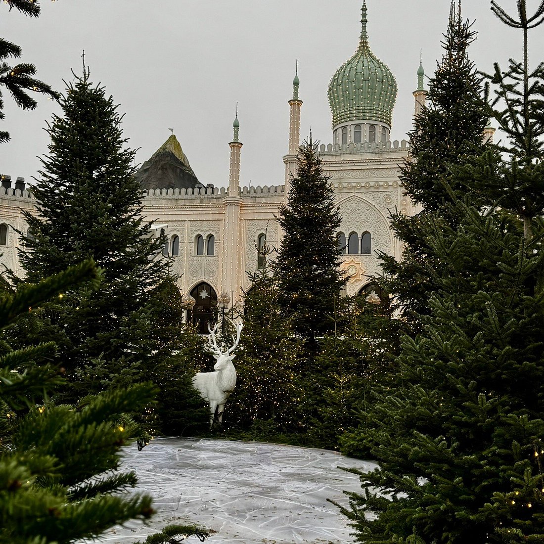 White reindeer statue on snowy path with evergreen trees and ornate palace with green domes in background