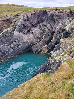 Rocky coastal cliff with turquoise water in cove, green grass above under cloudy sky