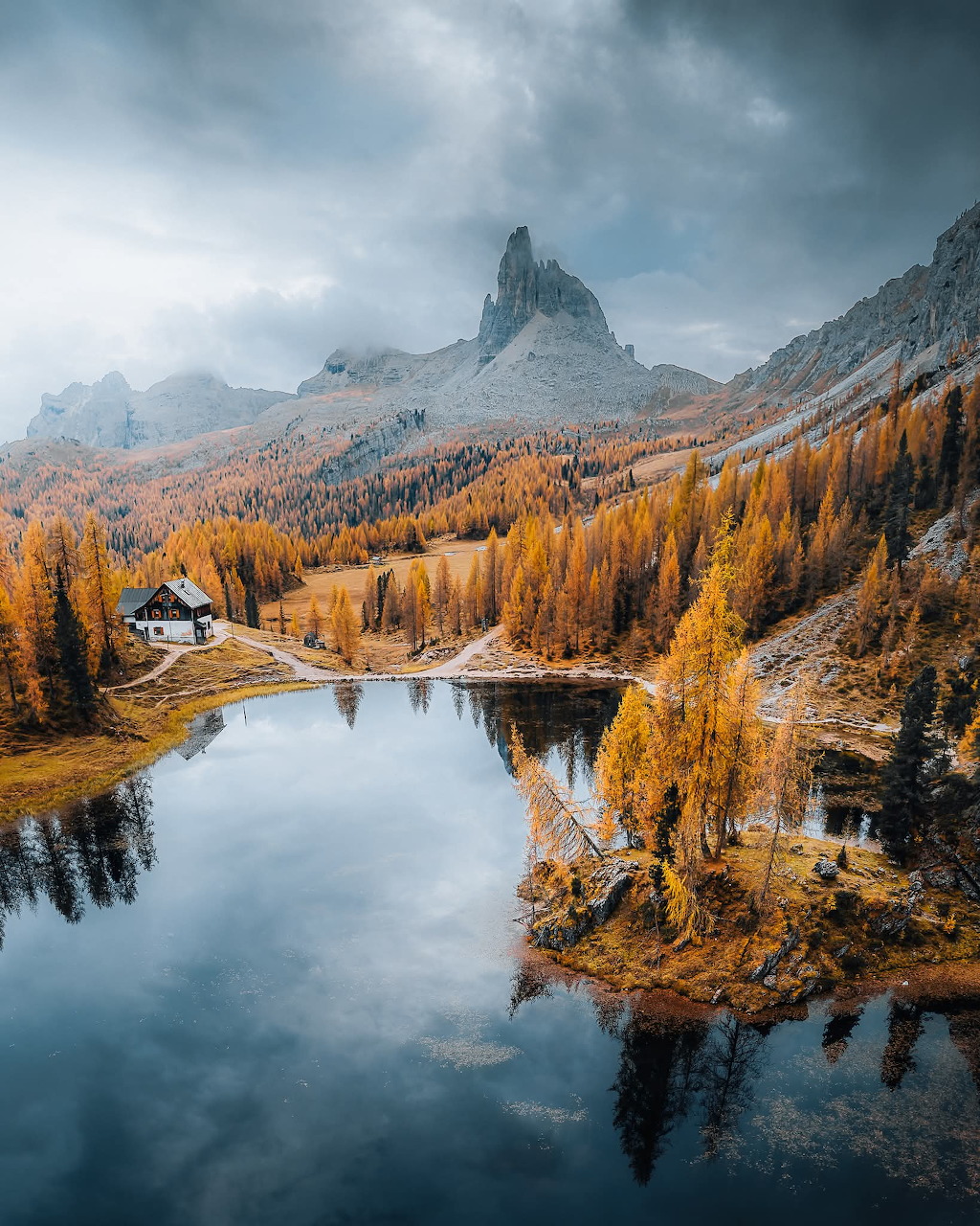 Aerial view of a mountain lake surrounded by golden autumn trees and rocky peaks with a small chalet