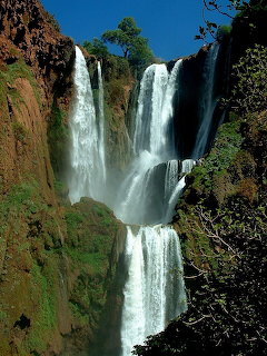 Multi-tiered waterfall on red-brown cliffs with trees and blue sky