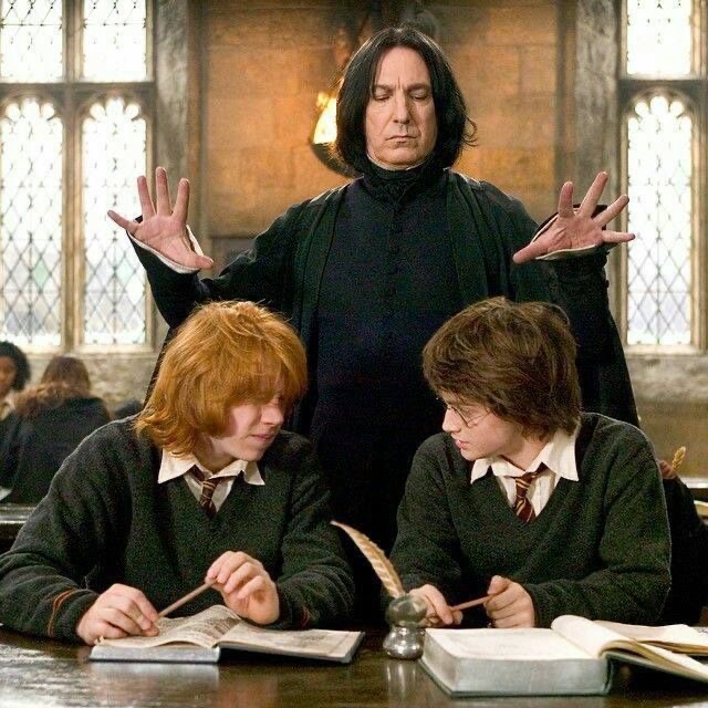 Dark-haired professor standing behind two students writing at a desk in a classroom