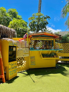 Yellow tropical food shack with coffee and fruit signs surrounded by palm trees and green lawn