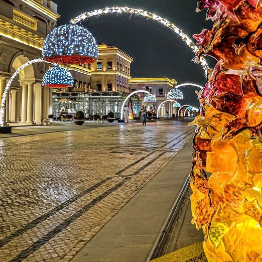 Night city street with decorative arch lights, ornate buildings, and a glowing orange sculpture