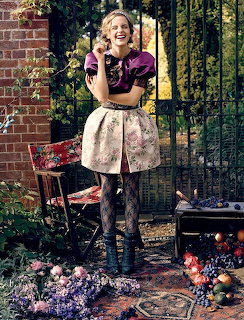 Emma Watson laughing in a garden wearing a purple top and floral skirt with patterned tights