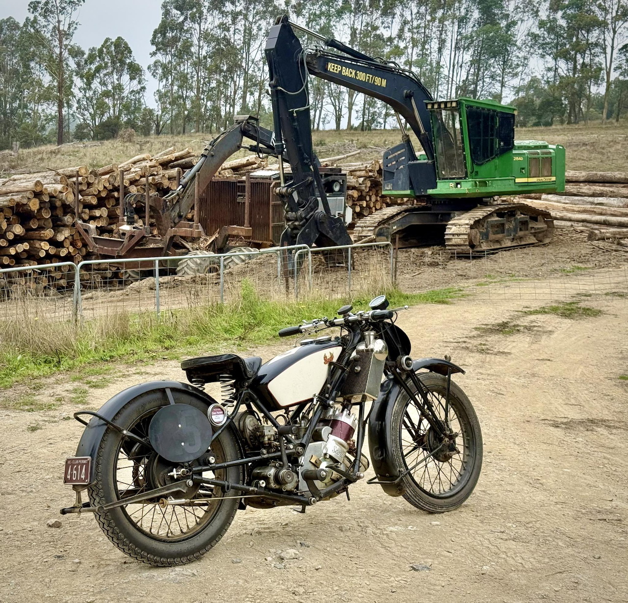 Classic black Scott motorcycle parked in front of green excavator moving logs at timber yard