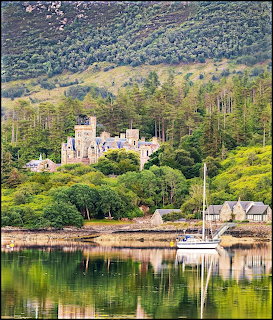 Stone castle surrounded by forest on loch shore with mountain backdrop and moored boats in foreground