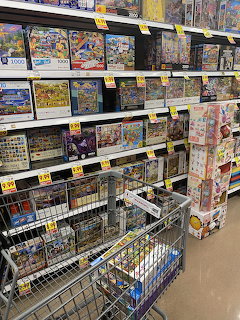 Store shelves stocked with colorful jigsaw puzzle boxes and yellow price tags with a shopping cart in foreground