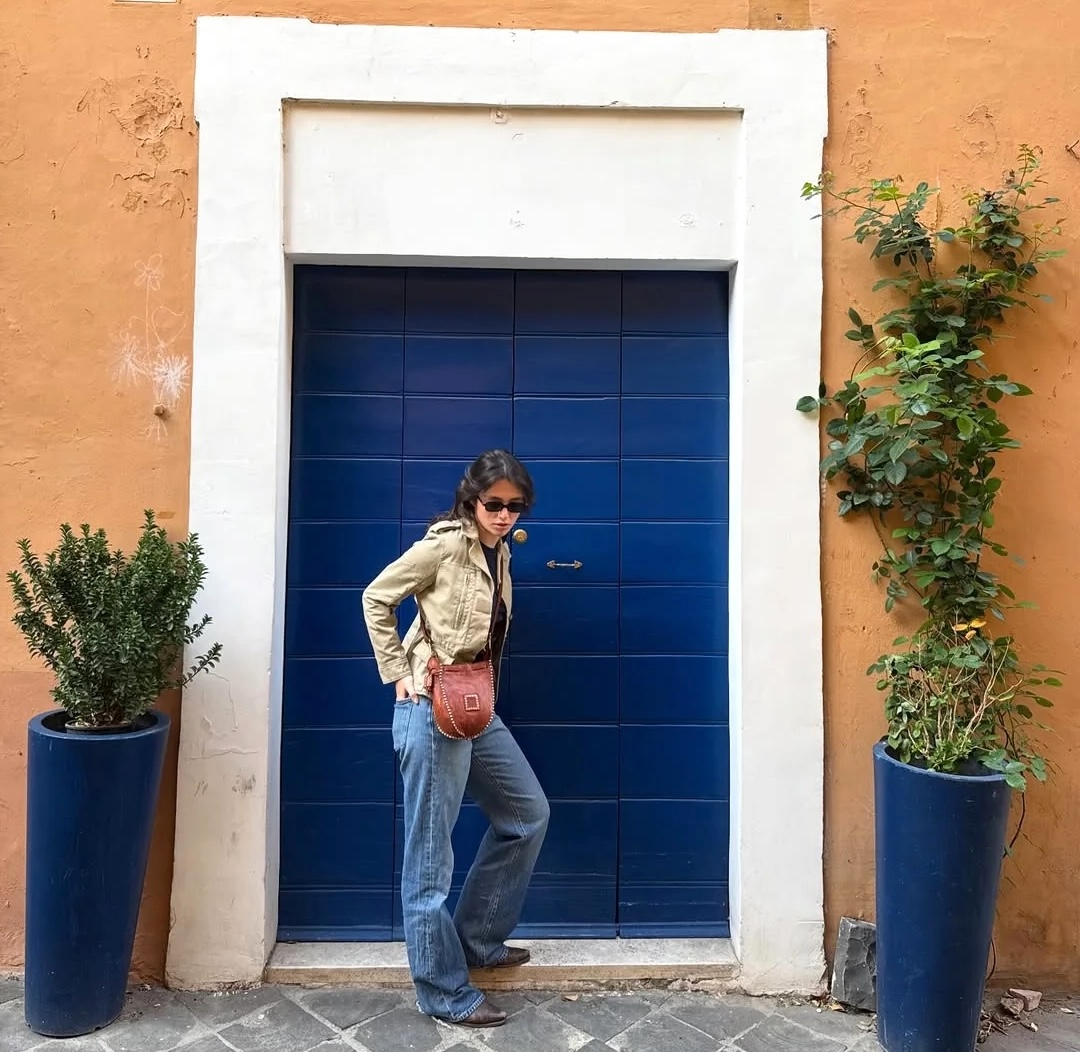 Woman in jeans and jacket posing by blue door with white frame on orange wall with plants