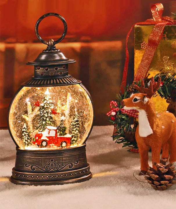 Illuminated Christmas snow globe with red truck and trees next to a reindeer figurine and gift box