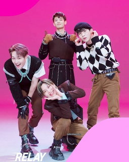 Four K-pop boy band members in casual streetwear posing on pink gradient background