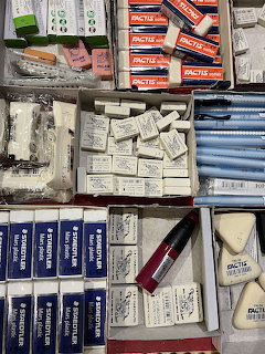 Open boxes of assorted stationery supplies on store shelves, including erasers, pens, and packaged items