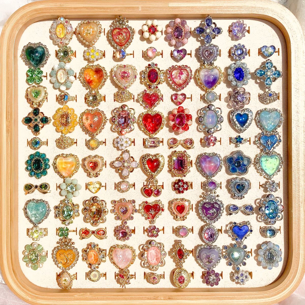 Grid of colorful gemstone rings with hearts and ornate settings displayed on white tray