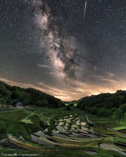 Milky Way arching over green terraced rice paddies at night with meteor streak and mountain silhouettes