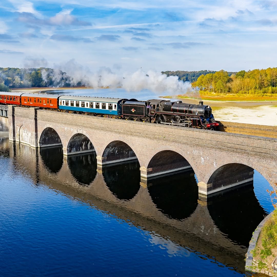 Vintage steam train crossing stone arched bridge over reflective water with autumn landscape