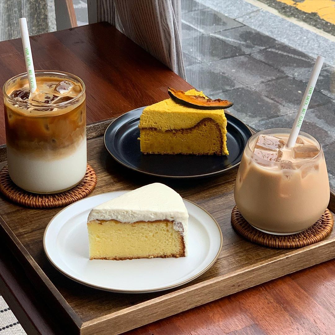 Two iced coffee drinks and two slices of cake on a wooden tray by a window