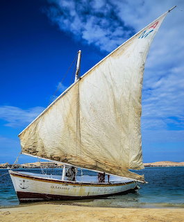 Traditional wooden sailboat with large white sail on calm turquoise water against blue cloudy sky