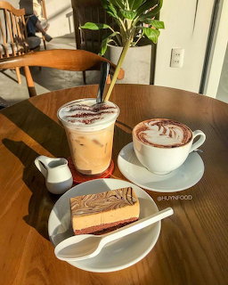 Two coffee drinks with latte art and a slice of cake on a wooden cafe table with plants in background
