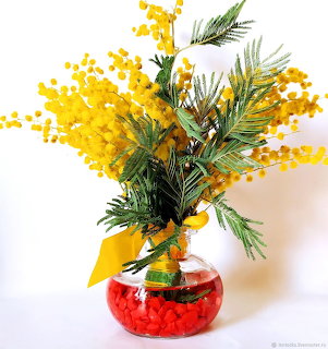Yellow mimosa flowers and green branches in a round glass vase with red pebbles