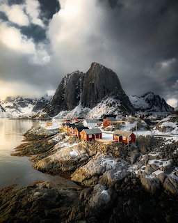 Red wooden cabins on snowy rocks below steep mountains in Hamnoy, Lofoten, Norway with dramatic clouds