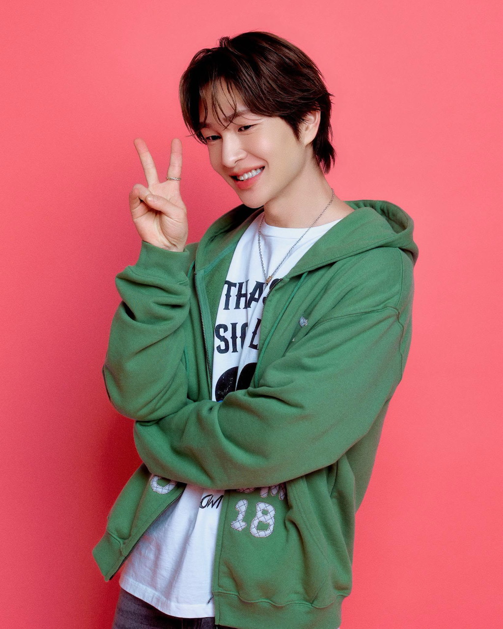 Smiling young man in a green hoodie making a peace sign in front of a pink background.