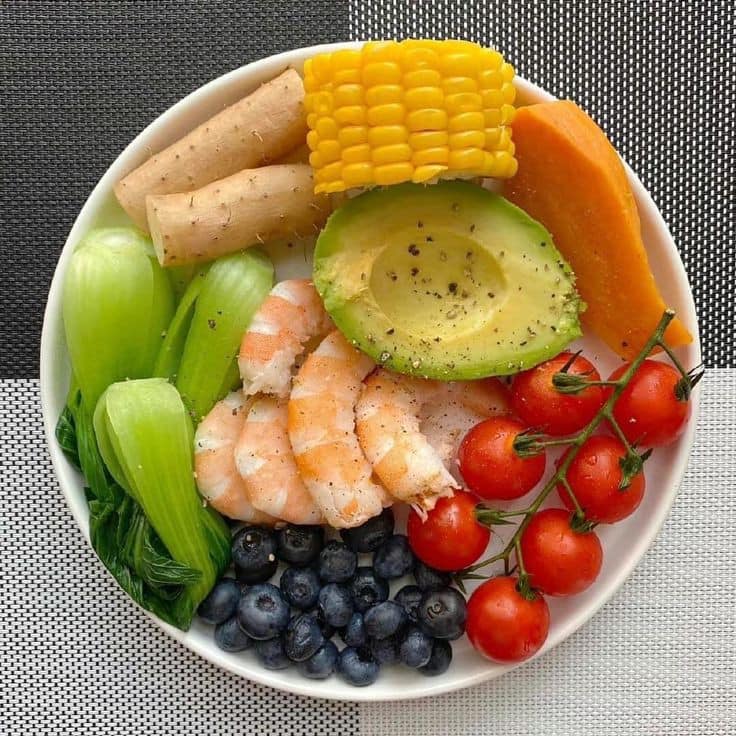 Colorful buddha bowl with shrimp, avocado, corn, tomatoes, blueberries, bok choy and breadsticks on gray background