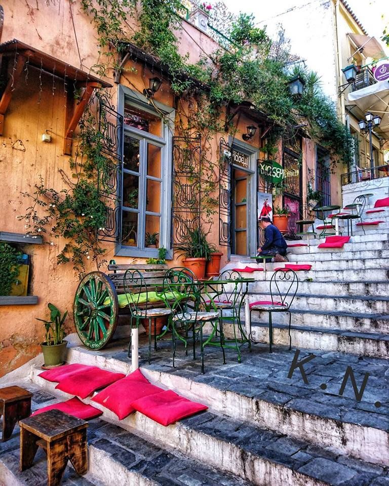 Outdoor cafe tables and colorful chairs on stone steps beside ochre building with climbing vines in Athens