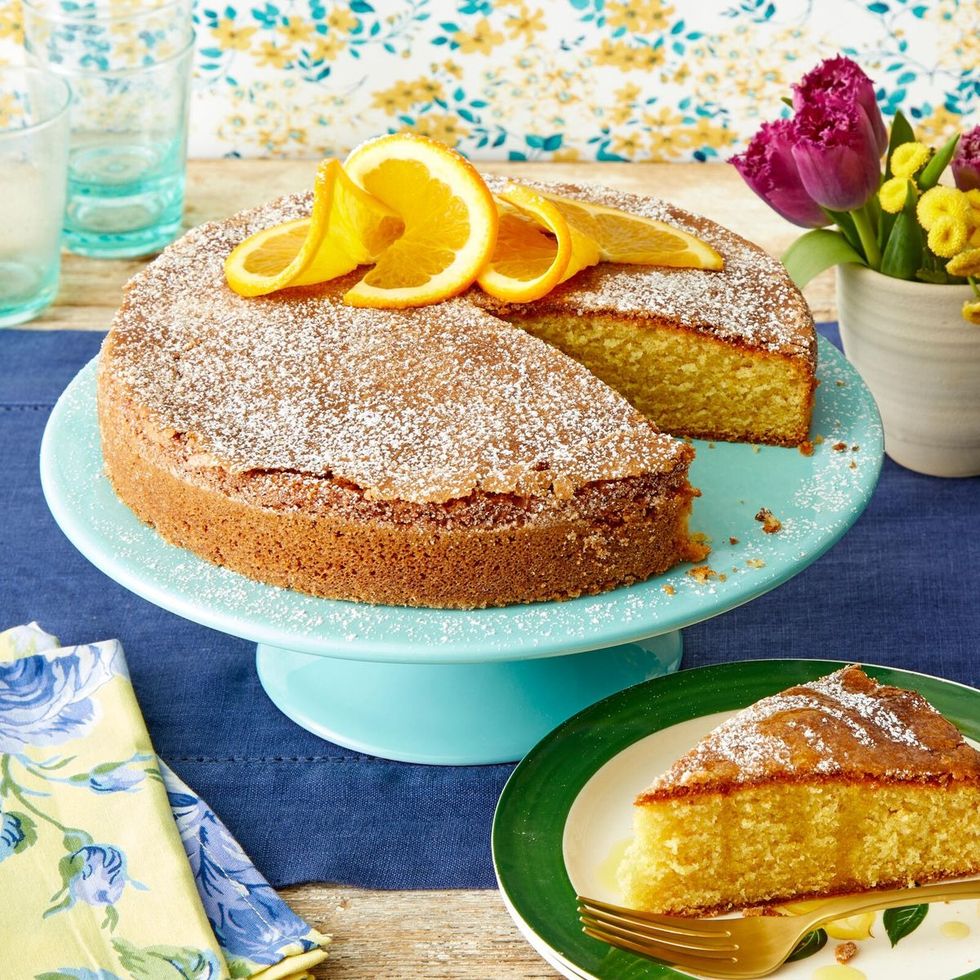 Lemon cake with powdered sugar and lemon slices on turquoise cake stand, slice on green plate, flowers in background