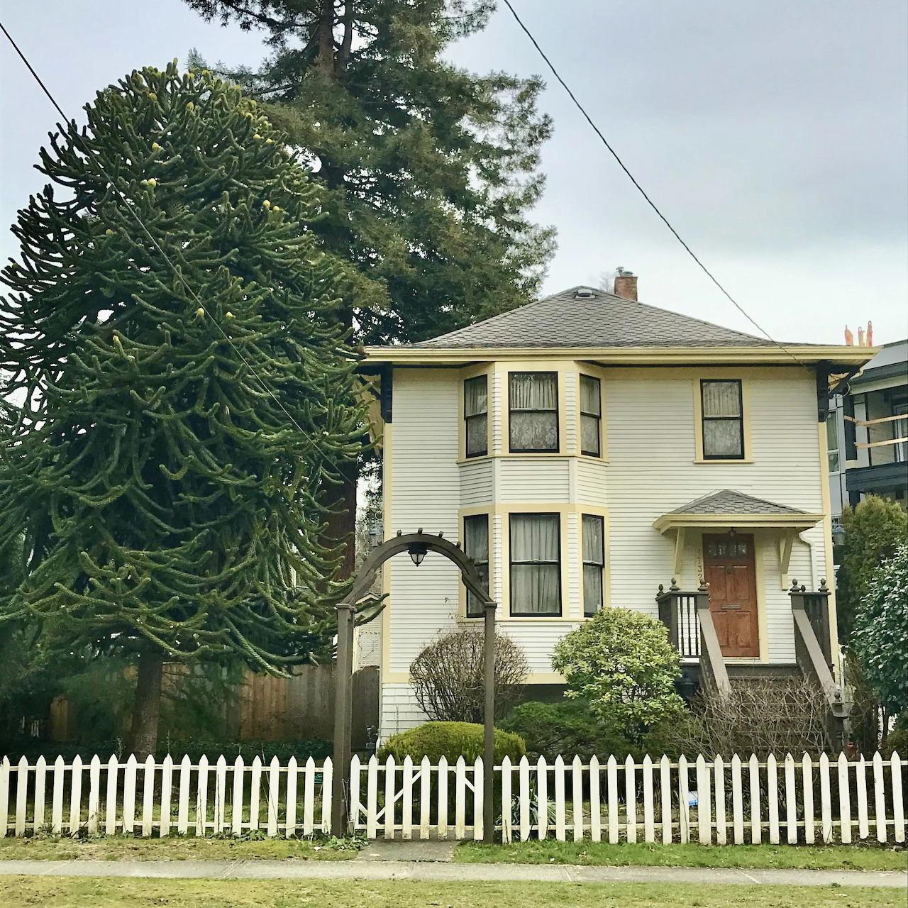 Cream two-story house behind a white picket fence with tall trees beside it