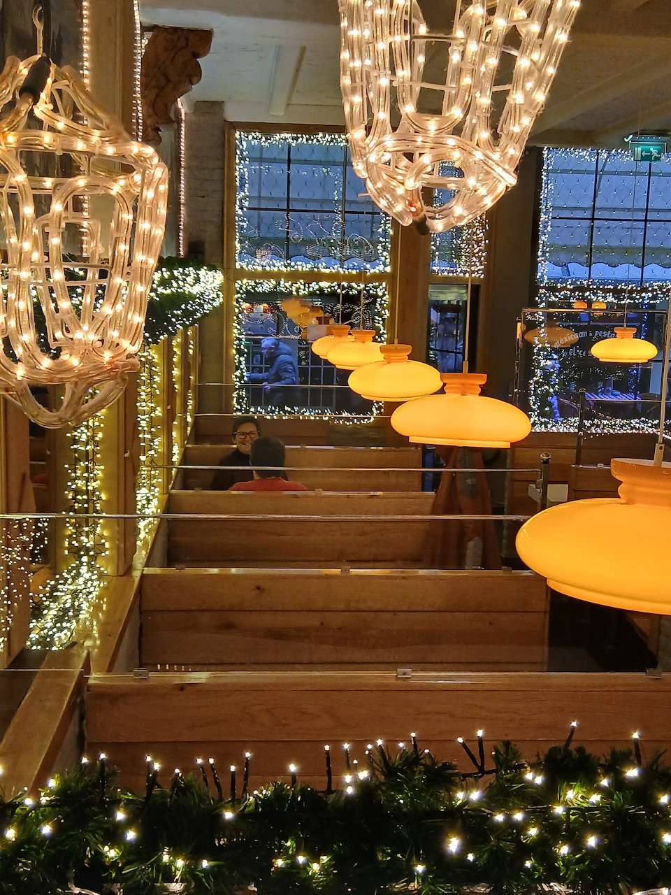 Restaurant interior with Christmas fairy lights, garlands, chandeliers, and orange pendant lamps over wooden tiered seating.