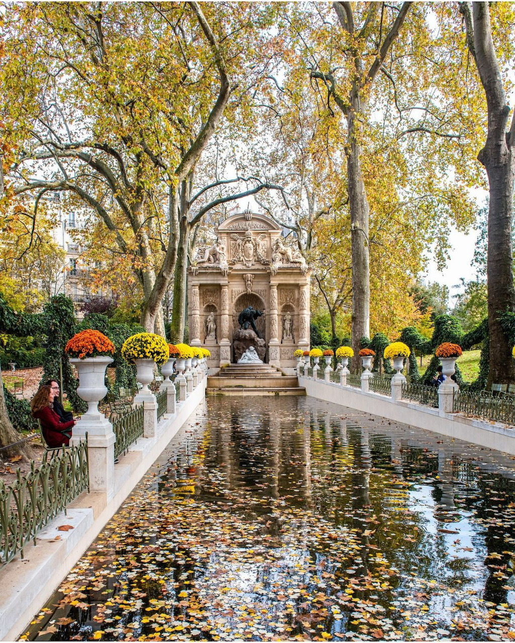 Ornate stone fountain with reflecting pool covered in fallen autumn leaves, surrounded by yellow trees in a park setting