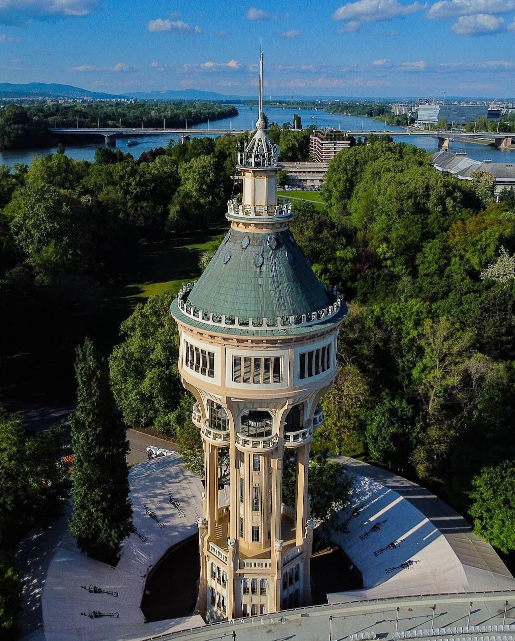Aerial photo of ornate beige water tower surrounded by trees on Margaret Island with river and cityscape behind
