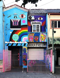 Colorful street art mural covering a small building facade with painted faces and eyes, pink gate in front