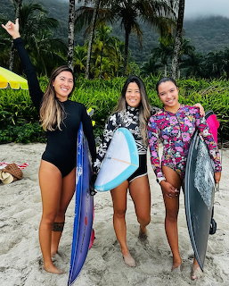 Three women surfers with surfboards on beach with palm trees and mountains behind