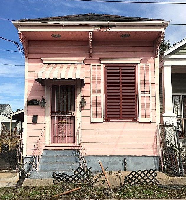 Small pink cottage with white trim, striped awning over door, dark shutters, and front yard