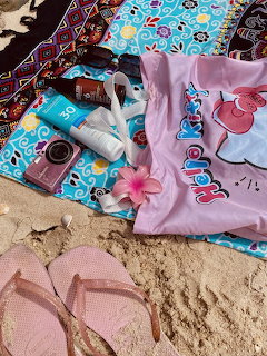Beach items including flipops, sunscreen, camera, Hello Kitty shirt, and flower on a towel on sand