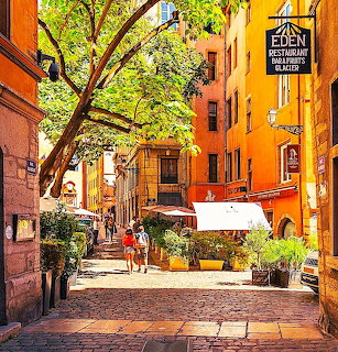 Sunny courtyard in Lyon with orange buildings, cobblestone pavement, trees, and people walking
