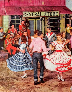 Square dancers in colorful dresses and western wear performing outside a red wooden general store building