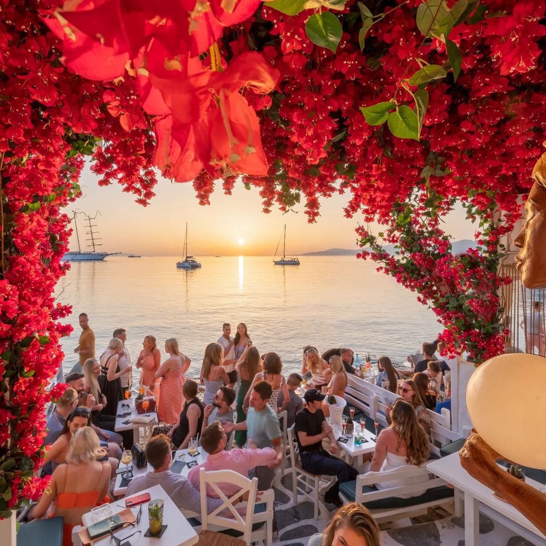 Outdoor restaurant terrace with red bougainvillea arch framing sunset ocean view and diners at tables