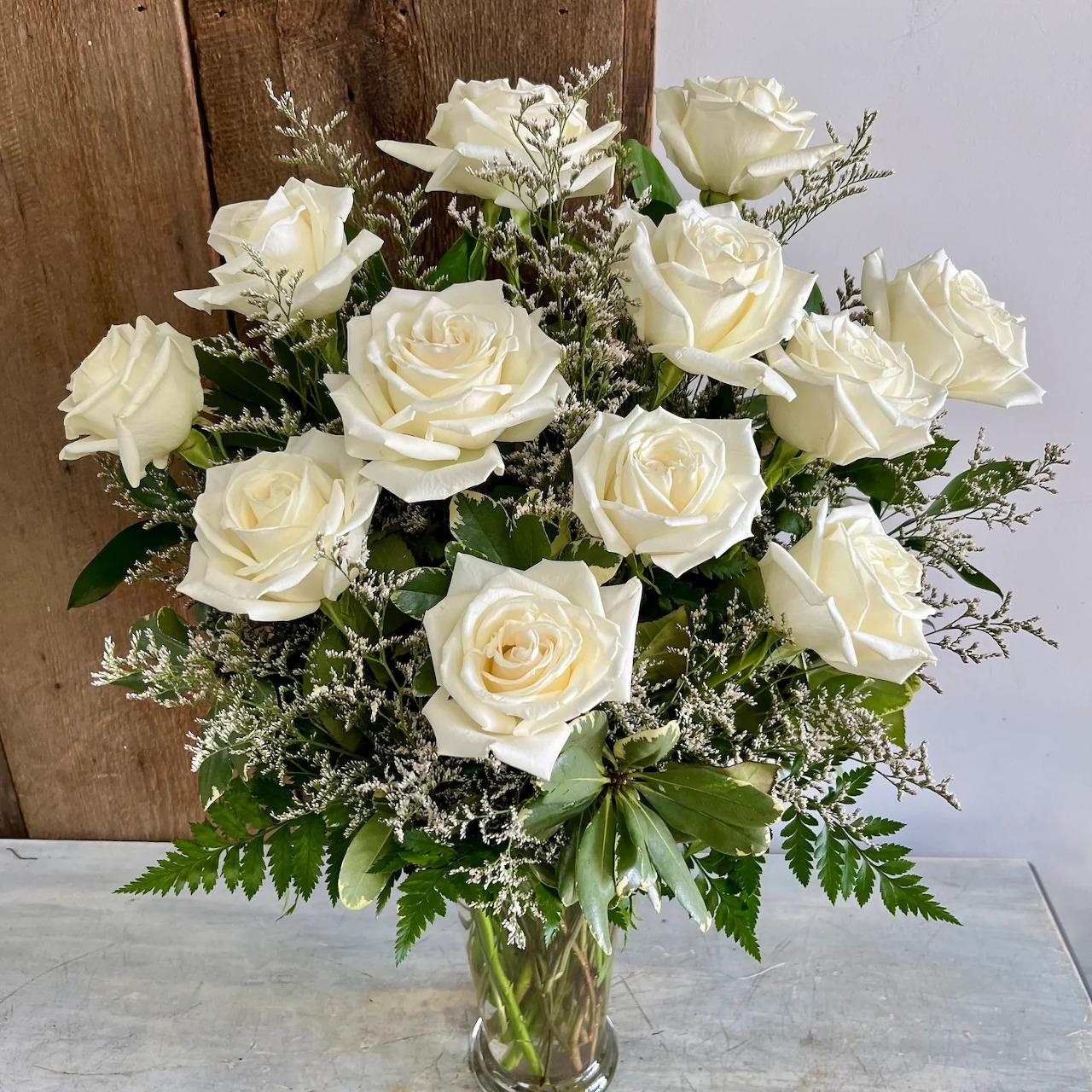 Bouquet of white roses with baby's breath and ferns in glass vase on white table with wood backdrop